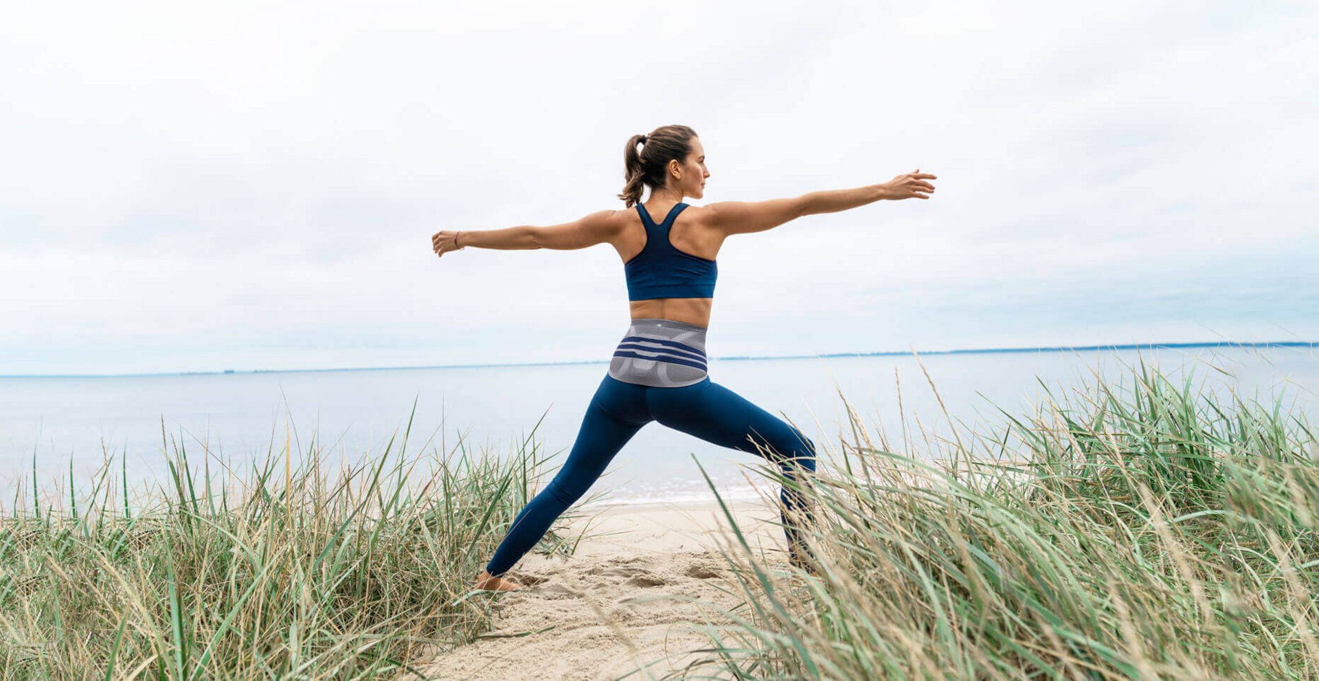 Das Bild zeigt eine junge Frau, die am Strand eine Yoga-Pose ausführt und dabei eine LumboTrain Lady Bandage zur Linderung von Rückenschmerzen trägt.