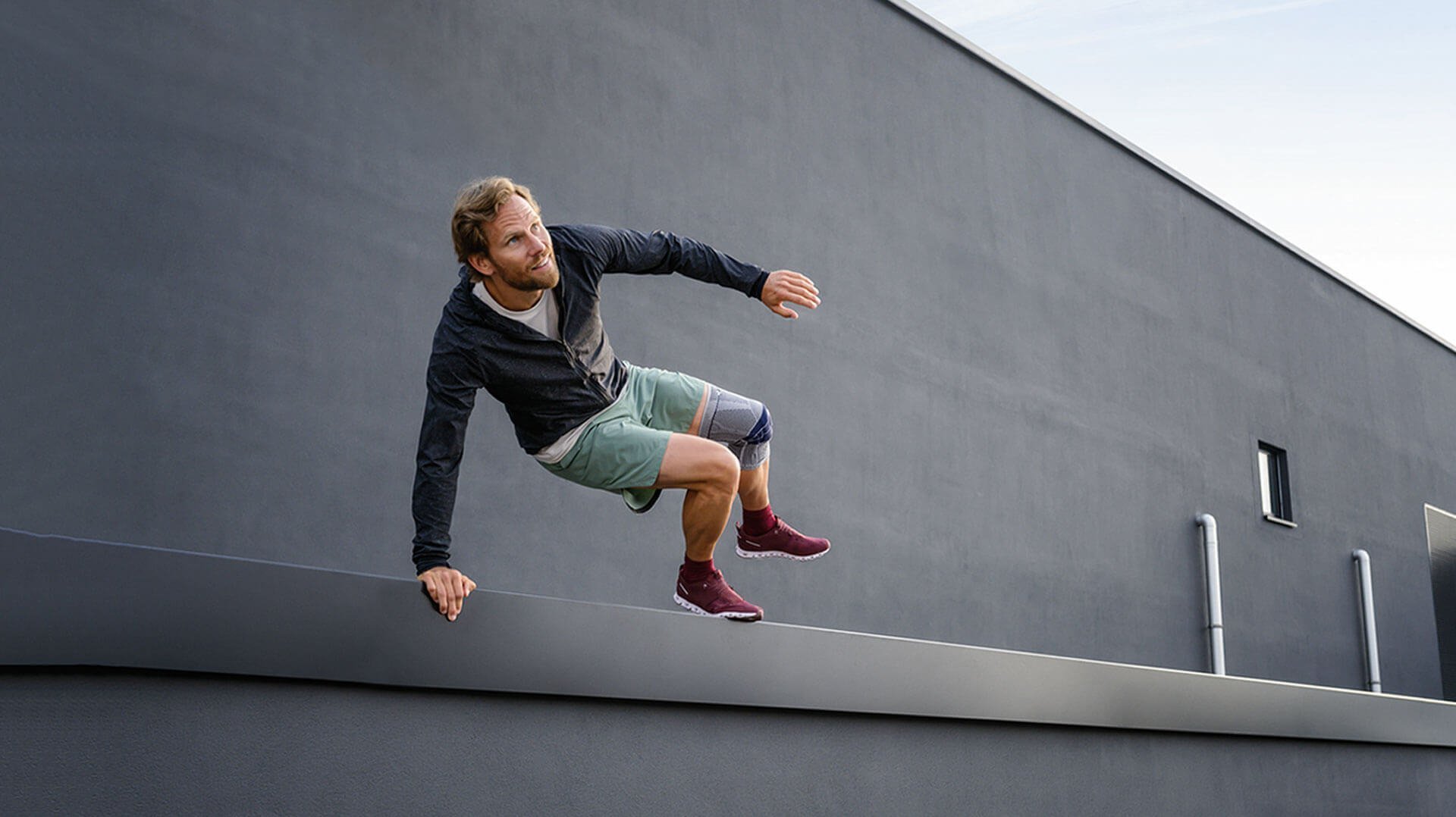 Abgebildet ist ein sportlicher Mann, der über ein Geländer vor einem Gebäude springt. Er trägt eine GenuTrian, eine schmerzlindernde Kniegelenksbandage von Bauerfeind, am linken Knie.